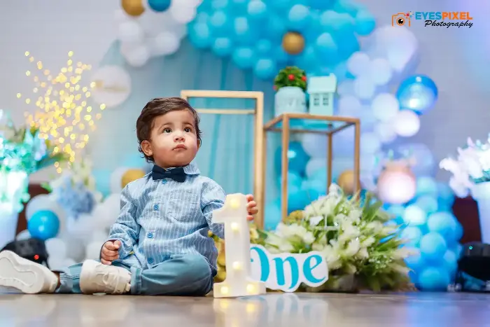 One-year-old boy in a blue shirt and bow tie beside a light-up ONE sign at a blue and gold balloon setup