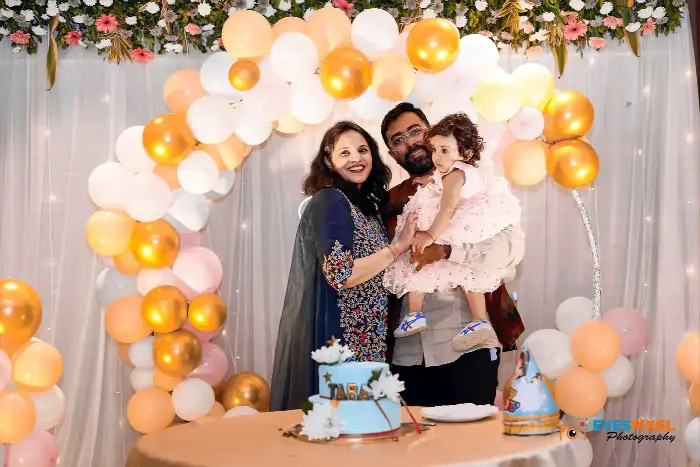 Parents holding their daughter beside her first birthday cake under a balloon arch
