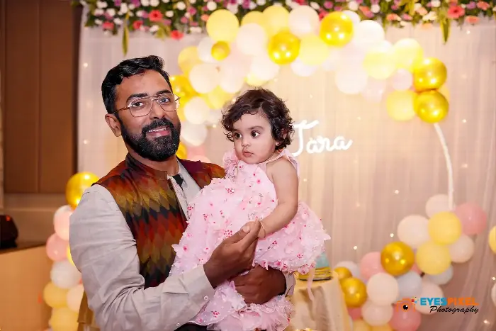 Father holding his daughter in front of a pink and yellow balloon arch with her name in lights