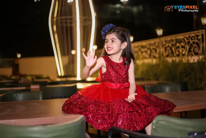 Young girl in a red sequinned party dress waving, seated at an outdoor evening venue