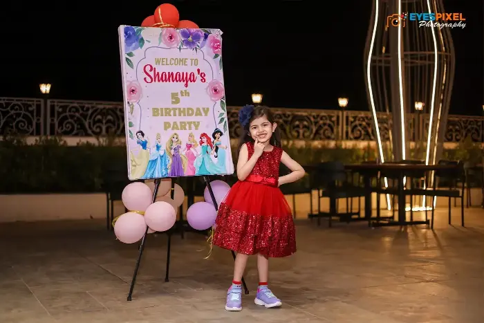 Girl in a red party dress beside a princess-themed fifth birthday welcome board