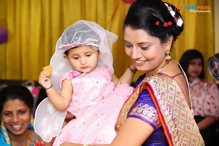 Mother in a purple saree holding her baby daughter in a veil at a birthday party