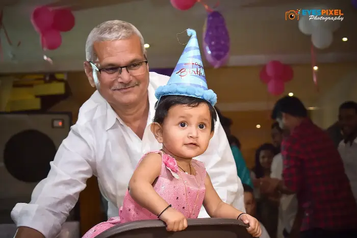Grandfather with a baby wearing a birthday hat at a party with balloons behind them