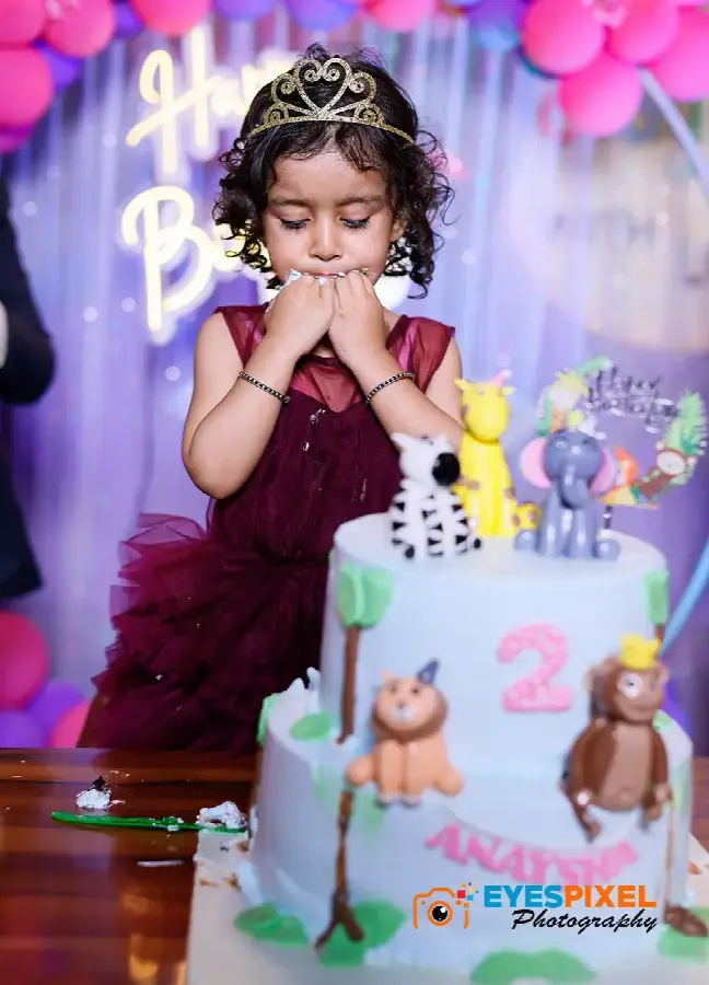 Girl in a tiara licking icing from her fingers beside her jungle-themed second birthday cake