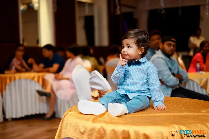 One-year-old in blue sitting on a banquet table during his birthday party, guests behind