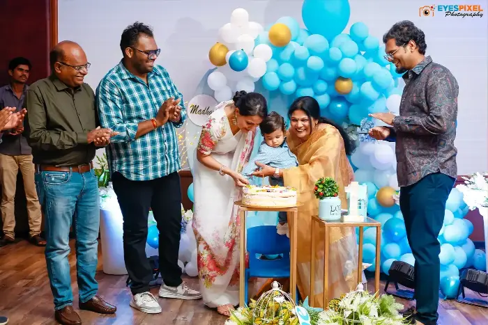 Family cutting the cake together at a first birthday party, guests applauding around them