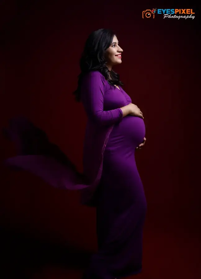 Side profile of an expectant mother in a purple gown cradling her bump against a dark red backdrop