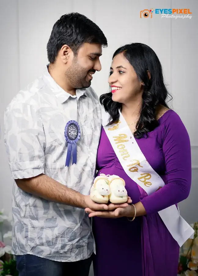 Expectant parents in Mom To Be and Dad To Be sashes holding a pair of baby shoes together