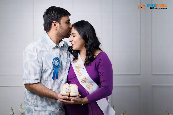 Father-to-be kissing the mother-to-be on the forehead as they hold baby shoes between them