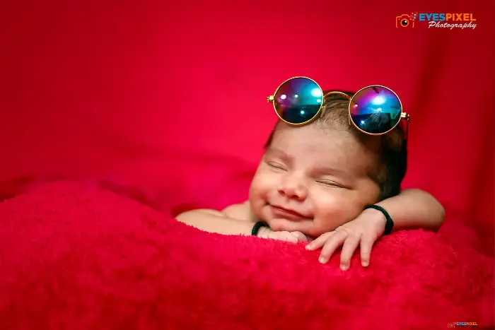 Smiling sleeping newborn on a red blanket with round prop sunglasses pushed up on their head, Pune