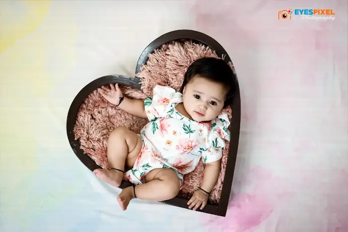 Baby a few months old lying in a heart-shaped wooden prop lined with pink fur, floral romper, Pune studio