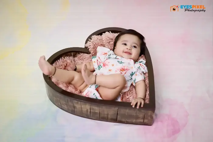 Smiling baby in a floral outfit propped in a heart-shaped wooden crate with pink fur, Pune