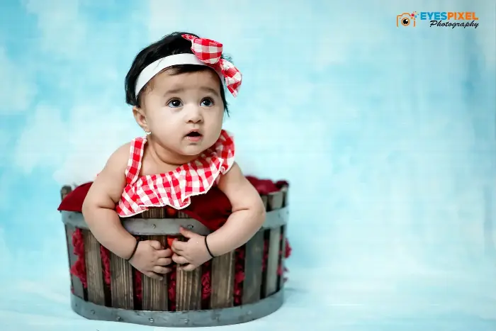 Baby girl in a red gingham outfit and matching headband, sitting in a wooden bucket against a blue backdrop