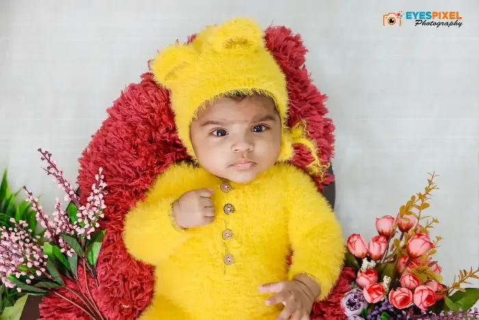 Baby in a yellow bear-eared knitted outfit against red fur and flowers, studio portrait in Pune