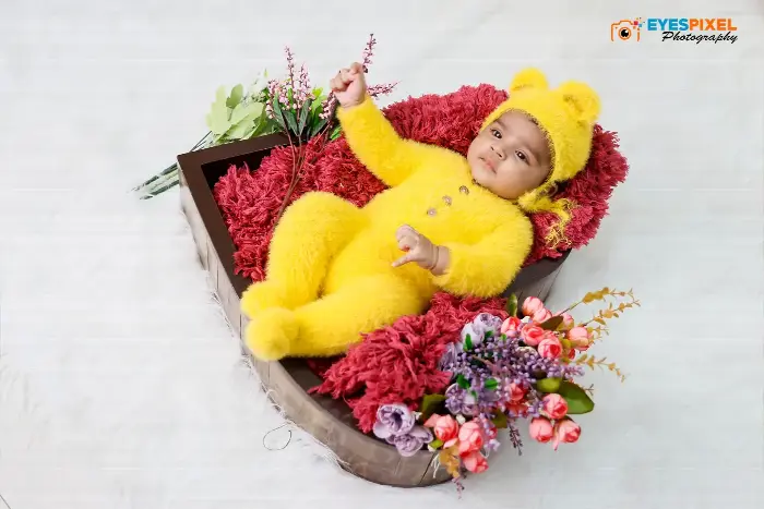 Baby in a yellow bear costume lying in a wooden crate with red fur and flowers, Pune studio