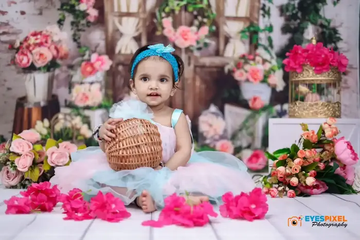 Toddler in a blue tutu holding a wicker basket on a floral studio set in Pune