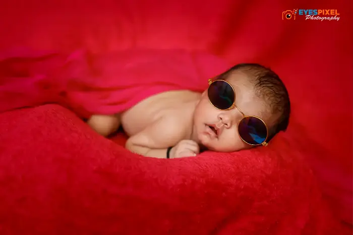 Newborn asleep on a red blanket wearing round prop sunglasses, photographed in Pune