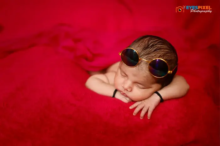 Newborn sleeping face down on folded red fabric with round prop sunglasses, Eyes Pixel studio, Pune