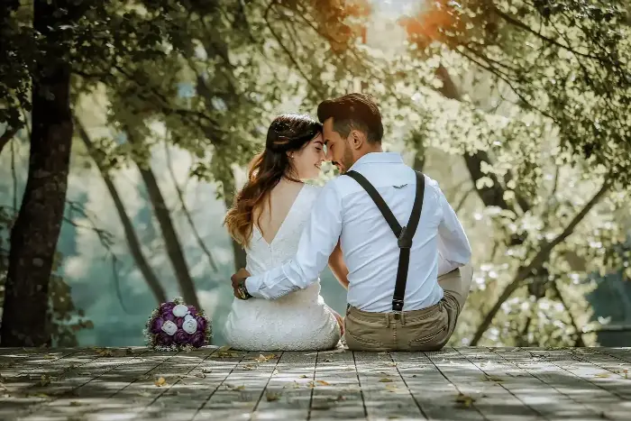 Couple seated on a paved terrace beneath sunlit trees, a bouquet on the ground beside them