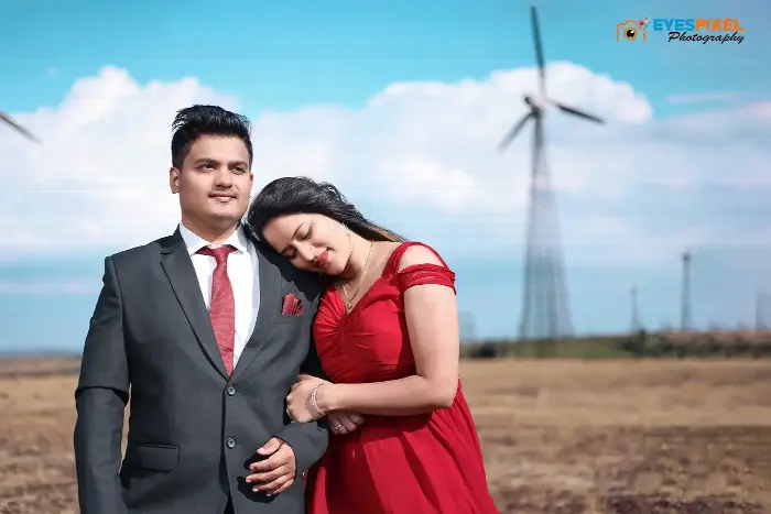 Woman in a red gown resting her head on her partner’s shoulder below wind turbines