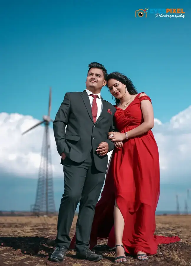 Couple in a grey suit and red gown standing on grassland below a wind turbine