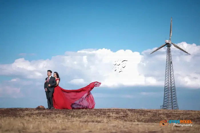 Couple small in the frame on open grassland, her red gown trailing, a wind turbine to one side