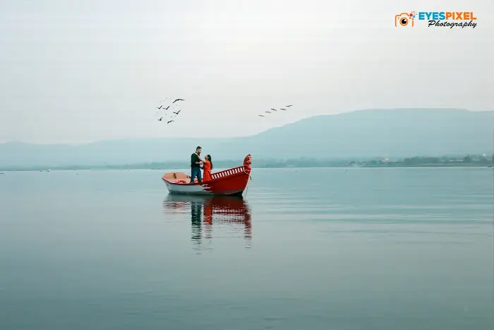 Wide view of a couple standing in a red boat on a lake, birds and hills beyond