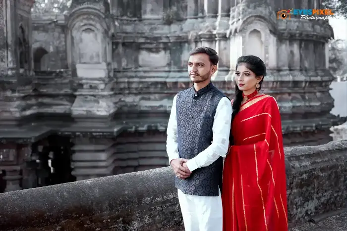 Couple standing hand in hand in front of an old stone temple, she in a red saree