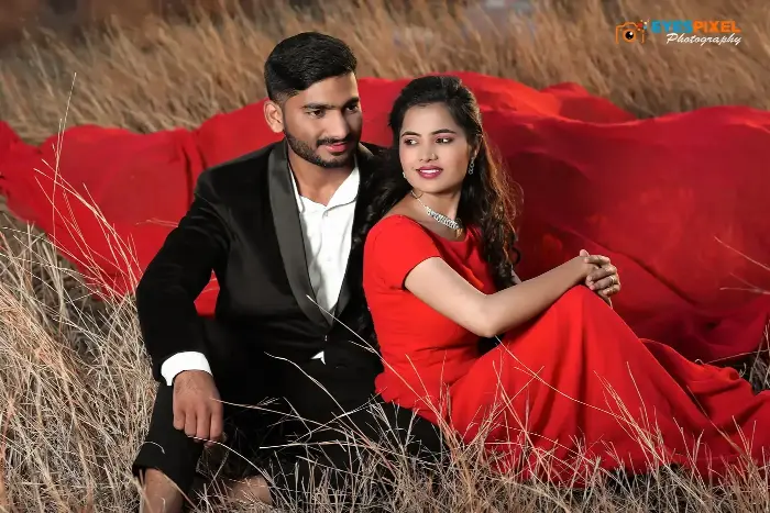 Couple seated in tall dry grass, she in a red saree with fabric trailing behind them