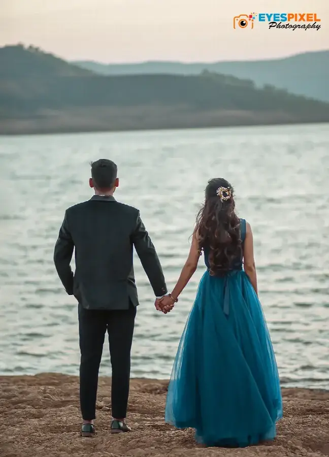 Couple seen from behind holding hands at the edge of a lake, hills across the water