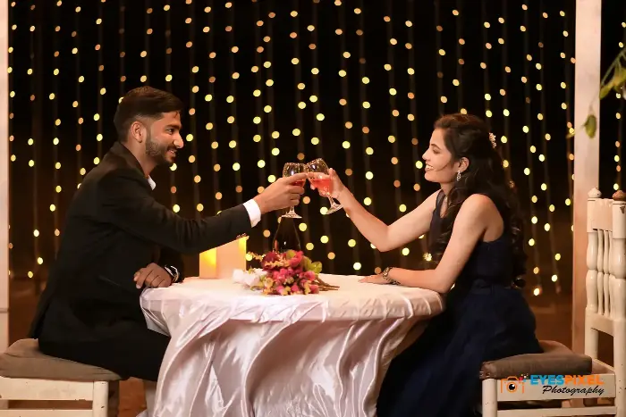 Couple raising glasses across a candlelit table against a wall of warm fairy lights