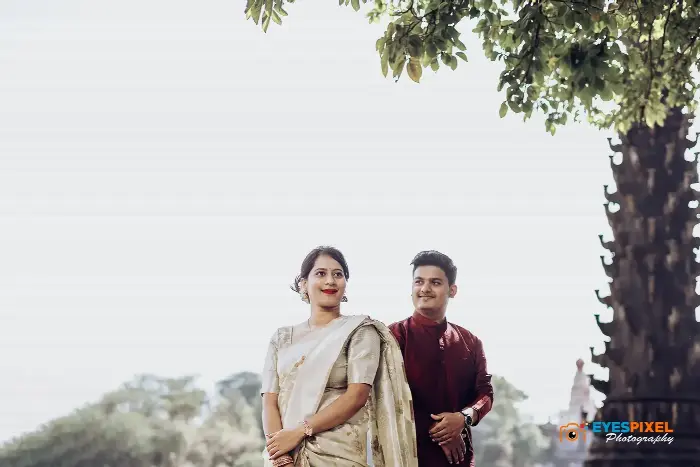 Couple in cream and maroon traditional dress standing under a tree near a temple