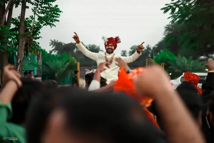 Groom in a red pheta with arms raised, carried through the wedding procession