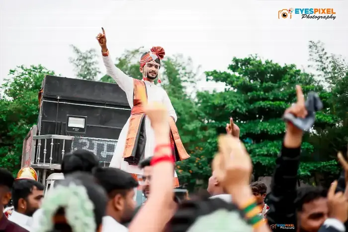Groom standing on a vehicle with a raised hand, crowd celebrating below him