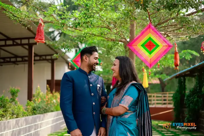 Couple in blue laughing beneath a tree hung with colourful kite decorations at a wedding venue