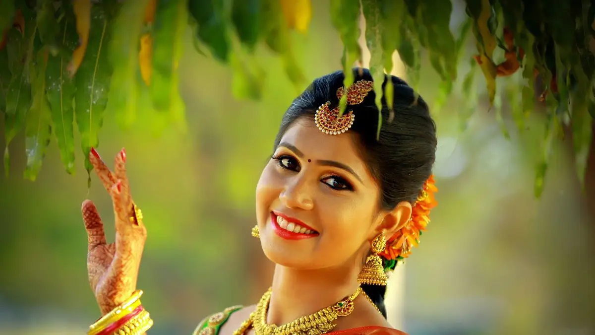 Bride in gold jewellery smiling beneath hanging leaves, marigolds in her hair